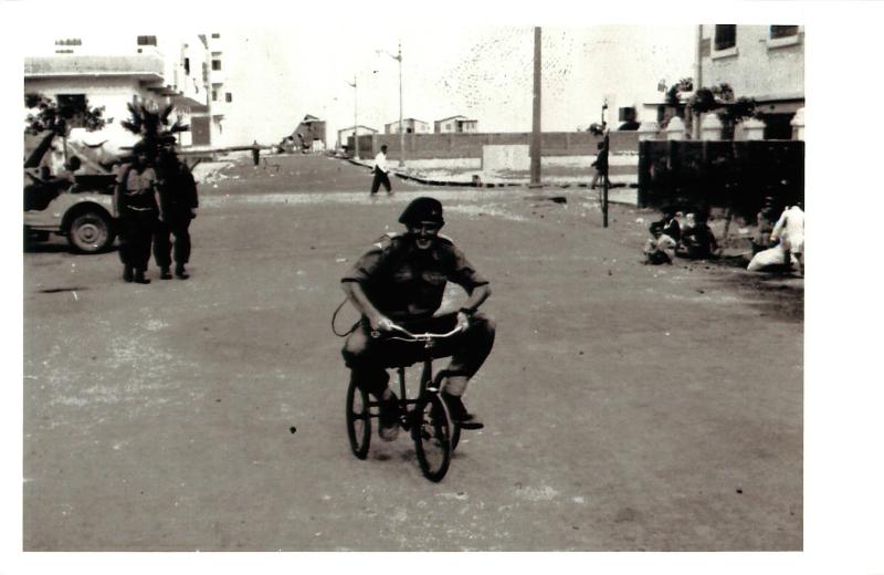 A smiling member of 3 PARA rides a small tricycle through a street in Port Said while locals look on.