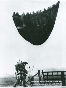 Under a parachute draped over telephone wires, a paratrooper carries a wounded man to aid.