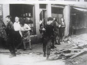 The aftermath of a bomb explosion. Paratroopers and civilians look nervously around amid debris.