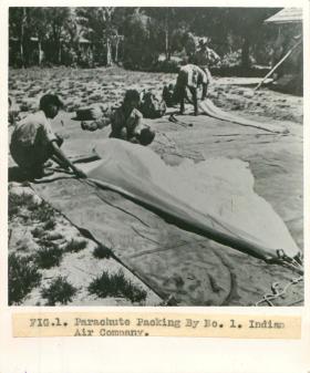 Men from No. 1 Indian Air Company squat on the floor outdoors and pack parachutes. 