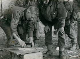 Paratrooper using a glass bottle to crush biscuits to make fishcakes. Others look on.  