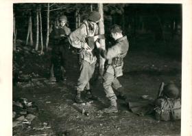 Three paratroopers stand in a wooded area at night. One helps the other with his kit. 