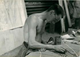 Bare-chested soldier works on Sicily briefing model with aid of aerial photo.
