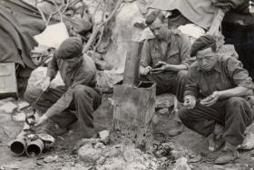 Privates Healy, Collins and Bowman crouch by a fire and make their cap badges at time of their introduction.