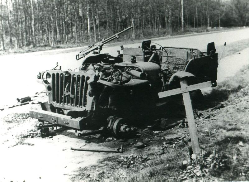 Burnt out Jeep on side of road. A wooden cross stands in teh foreground.