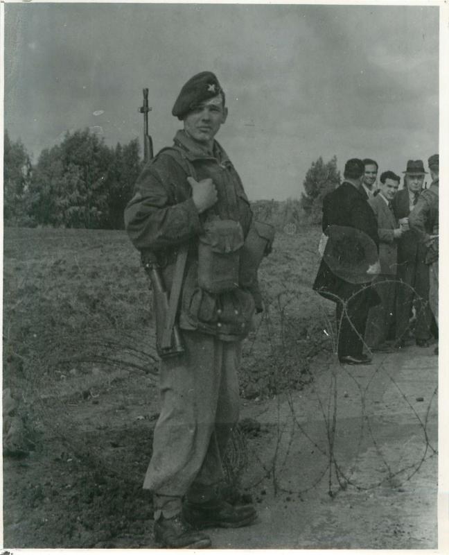 Paratrooper faces camera with barbed wire and civilians in the background. Palestine 1948.