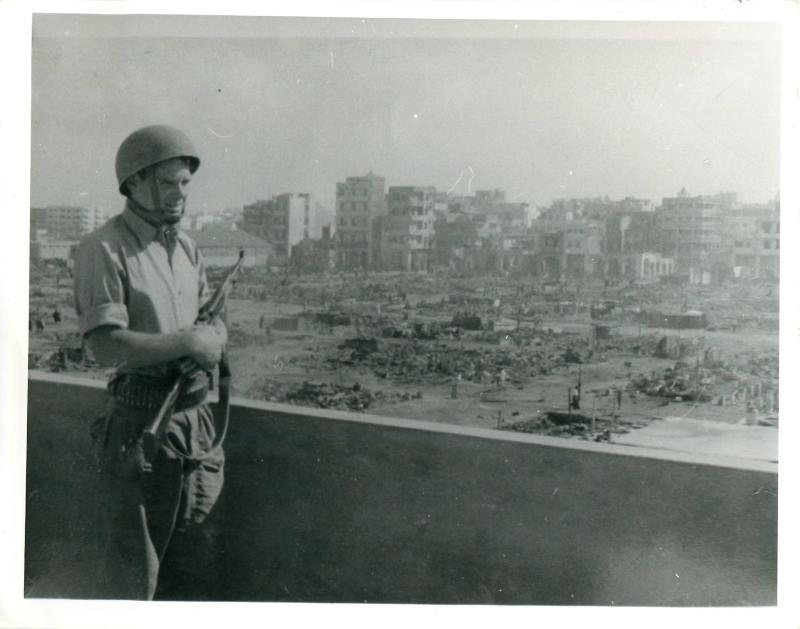 A member of 3 PARA stands on a balcony with a gun, overlooking a bomb-destroyed Port Said.