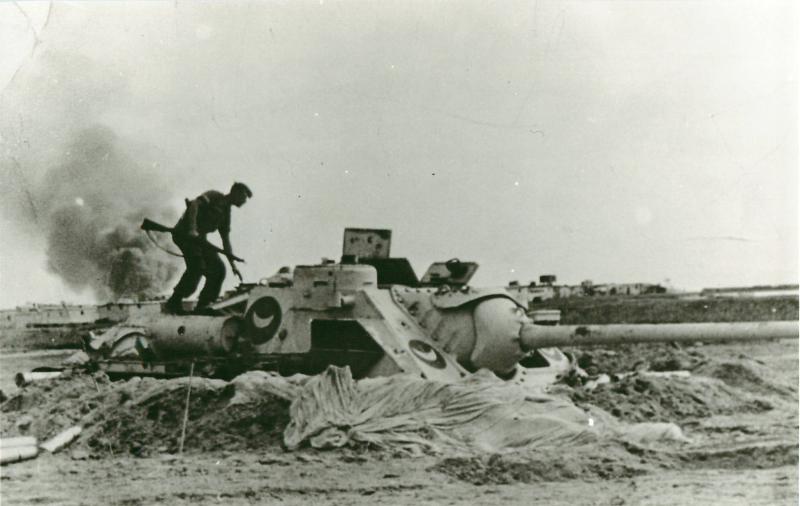 An SU-100 tank destroyer dug into sandy soil. A paratrooper stands on it holding a rifle.